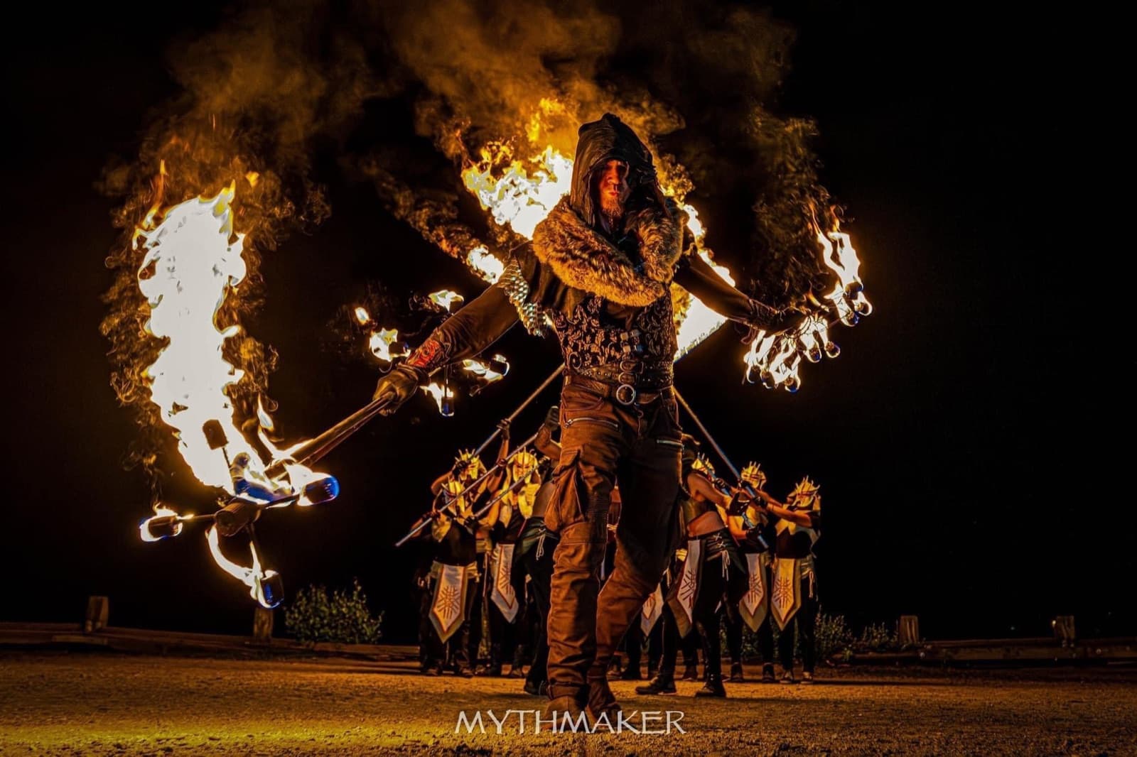 A hooded MythMaker fire dancer leads the troupe, twin arcs of flame trailing from his hands